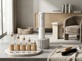 A white and clear personal care set with wooden accents, including a tall cylindrical device and four spray bottles on a marble tray, styled in a bright, modern minimalist room.