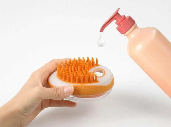 A hand holds an orange and white round scalp massager brush while a bottle of clear liquid with a pink pump is shown dispensing droplets above it on a white background.