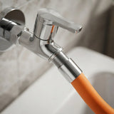 A close-up of a chrome wall-mounted faucet with a flexible orange hose adapter attached to the supply pipe, set against a blurred bathroom background.