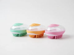 Three oval-shaped handheld scrubber brushes with clear domed tops and colorful bristle bases in mint green, orange, and pink, arranged on a white background.