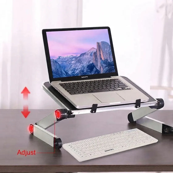 A silver aluminum laptop desk stand holding an open silver laptop, shown on a dark desk with a white keyboard in front. Red arrows and the word 'Adjust' indicate the stand's height-changing legs.