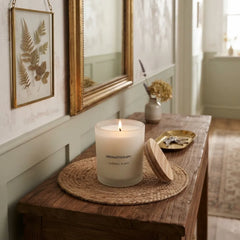 A lit white candle in a frosted glass jar sits on a woven placemat on a rustic wooden table, with its wooden lid leaning beside it. The setting is a styled interior with a mirror, framed botanical art, and a small vase in the background.