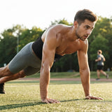 A shirtless man in gray athletic shorts and a black tank top performs a push-up on a grassy field outdoors, with trees and another runner blurred in the background.