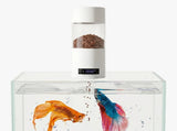 A white automatic fish food dispenser with a digital timer and clear container filled with brown flakes, placed on top of a glass aquarium containing two colorful betta fish.