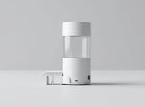 A minimalist white and clear cylindrical device with a small white accessory placed beside it, photographed on a clean light gray studio surface and background.