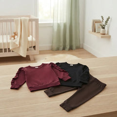 Three children's long-sleeve sweatshirt and jogger set pieces are laid out on a light wood floor in a softly styled nursery. The sets are in burgundy, black, and brown, each with ruffled sleeves and matching joggers.