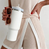 A close-up lifestyle shot of a person wearing a beige ribbed knit wrap dress with white contrast trim, holding a white travel tumbler and wearing rose gold-framed sunglasses.