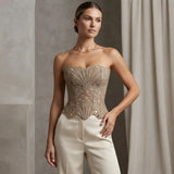 A woman models a strapless beige corset top with intricate floral embroidery and cutout detailing, paired with cream-colored trousers, set against a neutral studio backdrop with draped curtains.