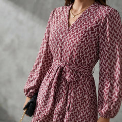 A woman wearing a long-sleeve wrap-style dress with a tie waist and an allover geometric pattern in dusty pink and white. She is holding a small black clutch with a gold chain strap against a textured gray background.