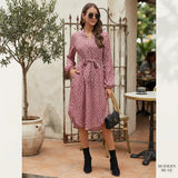 A woman models a pink and white floral print wrap dress with a tie waist, long sleeves, and a V-neckline in an outdoor patio setting. She is wearing black ankle boots, sunglasses, and holding a small black handbag.