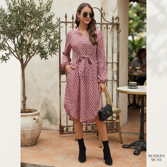 A woman models a pink and white floral print wrap dress with a tie waist, long sleeves, and a V-neckline in an outdoor patio setting. She is wearing black ankle boots, sunglasses, and holding a small black handbag.