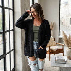 A woman wearing a black cable-knit open-front cardigan over an olive green tank top and distressed light-wash denim shorts, standing in a modern room with large windows.