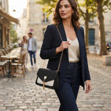 A woman walking on a cobblestone street wearing a navy blazer, cream camisole, and dark jeans, carrying a black and white flap shoulder bag with a gold chain strap.