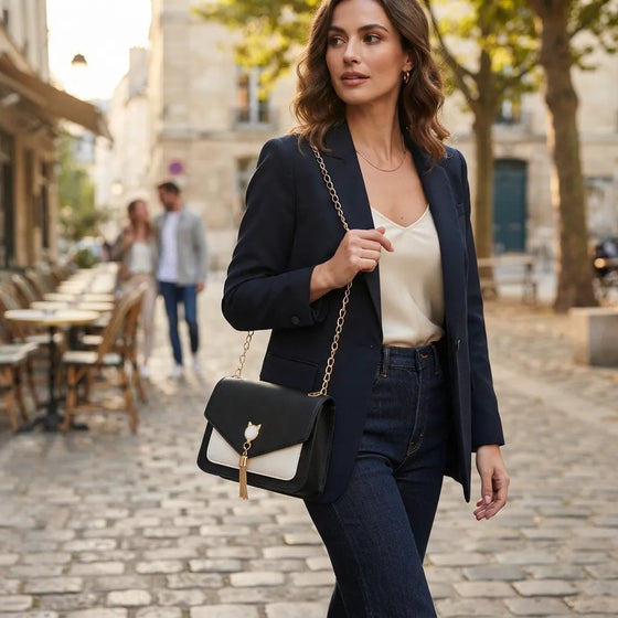 A woman walking on a cobblestone street wearing a navy blazer, cream camisole, and dark jeans, carrying a black and white flap shoulder bag with a gold chain strap.