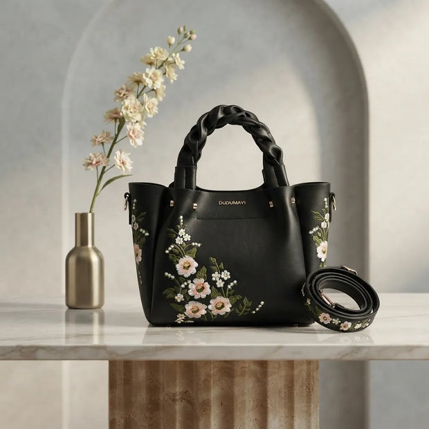 A black leather handbag with floral embroidery and braided top handles sits on a marble surface next to a gold vase with white flowers. A matching floral belt is coiled beside the bag, set against a modern gray arched wall.