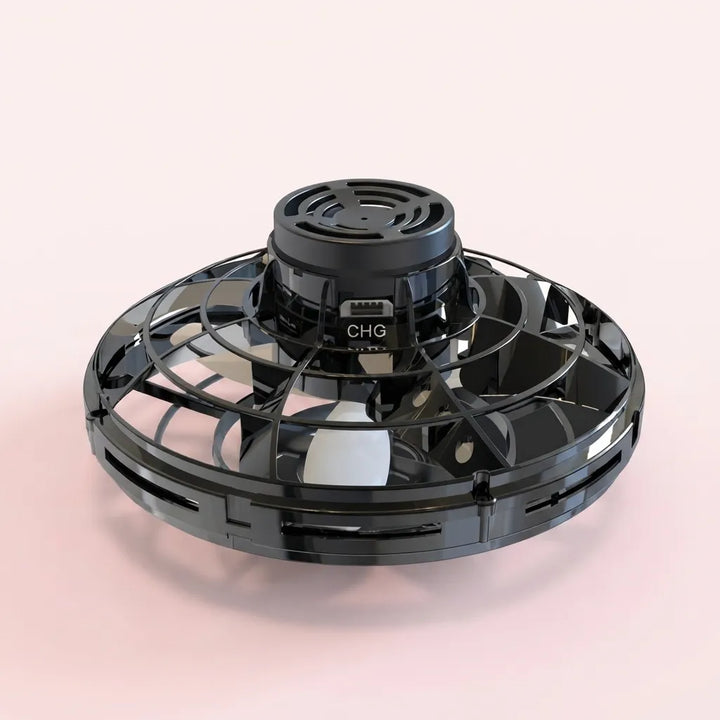 A black circular fan or vent-like appliance with a central motor housing and protective grille, shown on a soft pale pink studio background.