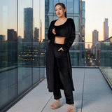 A woman stands on a modern glass balcony at sunset wearing a black two-piece lounge set consisting of a cropped long-sleeve top and matching pants, with a long open-front black cardigan draped over one shoulder. The background shows a city skyline at dusk.