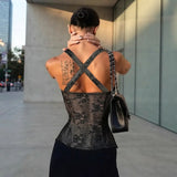 A woman photographed from behind wearing a black lace corset-style top with wide crisscross shoulder straps, standing outdoors near a modern glass building. She is carrying a black shoulder bag with a chain strap.