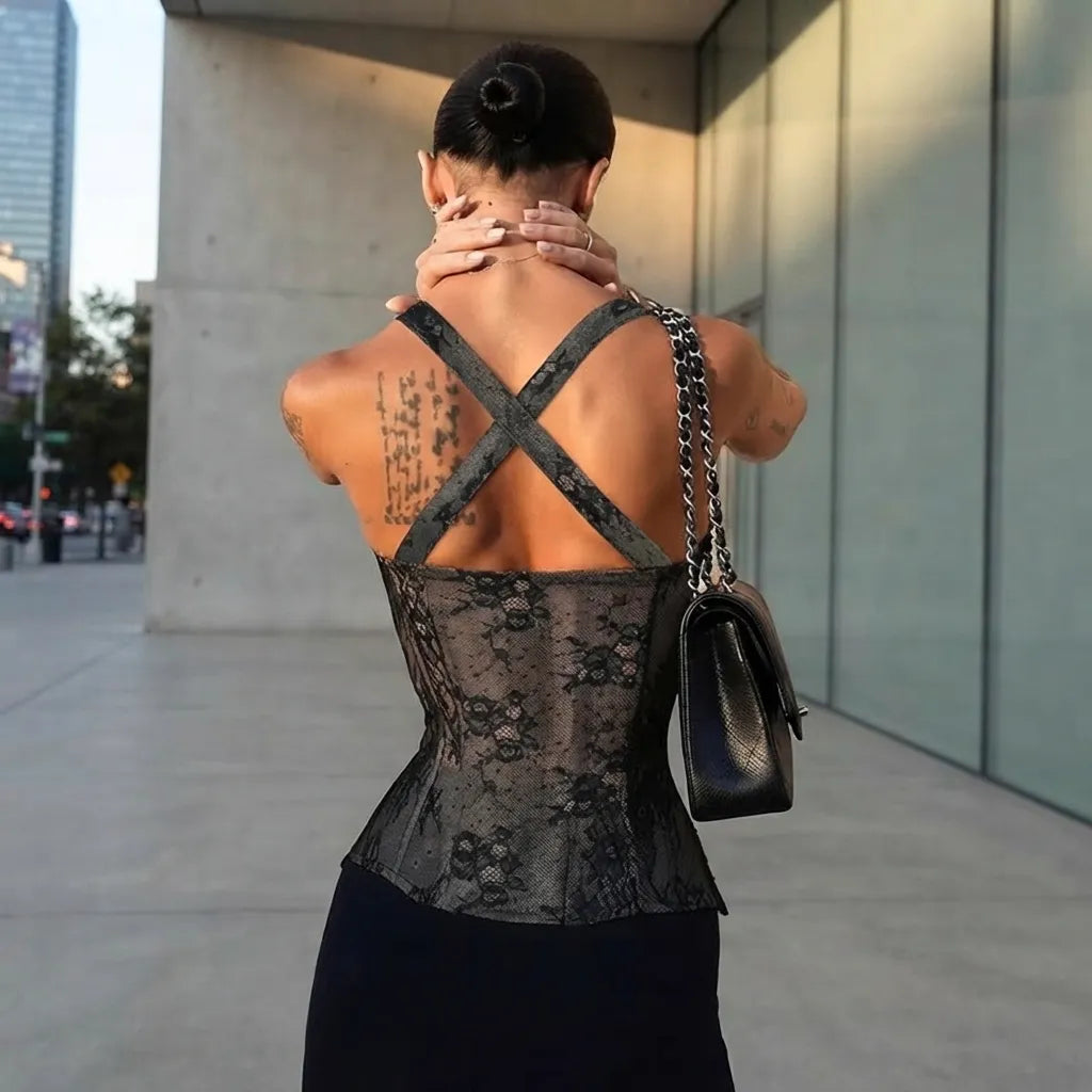 A woman photographed from behind wearing a black lace corset-style top with wide crisscross shoulder straps, standing outdoors near a modern glass building. She is carrying a black shoulder bag with a chain strap.