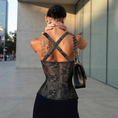 A woman photographed from behind wearing a black lace corset-style top with wide crisscross shoulder straps, standing outdoors near a modern glass building. She is carrying a black shoulder bag with a chain strap.