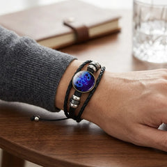 A close-up of a person's wrist wearing a multi-strand black leather bracelet with a round blue and purple cat face charm and silver-toned metal beads, resting on a wooden table.