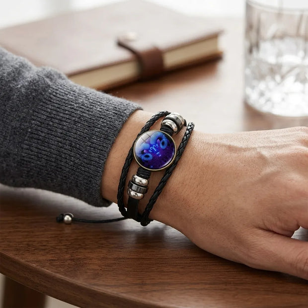 A close-up of a person's wrist wearing a multi-strand black leather bracelet with a round blue and purple cat face charm and silver-toned metal beads, resting on a wooden table.