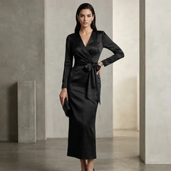 A woman models a black satin long-sleeve wrap midi dress with a tie waist, standing in a minimalist studio with concrete pillars.