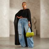 A model stands in a minimalist studio wearing a black sheer long-sleeve top, light-wash distressed wide-leg jeans, and holding a bright lime green textured shoulder bag.