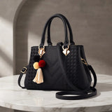 A black woven leather handbag with double top handles, a detachable shoulder strap, and decorative gold-tone hardware, including a tassel charm with red, yellow, and cream tassels. The bag is displayed on a white marble table against a neutral gray stone wall background.