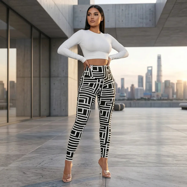 A woman stands on a rooftop terrace wearing black and white geometric patterned leggings, a white long-sleeve crop top, and clear high-heeled sandals, with a city skyline in the background.