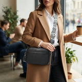 A woman in a camel coat and white shirt carries a black crossbody bag with a black-and-white patterned strap in a bright cafe setting.