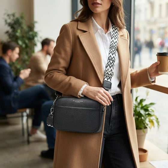 A woman in a camel coat and white shirt carries a black crossbody bag with a black-and-white patterned strap in a bright cafe setting.