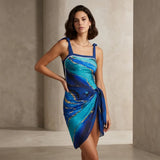 A woman models a sleeveless wrap-style dress with a blue and turquoise abstract print and gold veining, set against a neutral concrete-textured studio background.