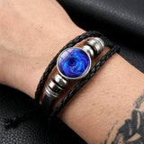 A close-up of a person's wrist wearing a multi-strand black leather bracelet with a central oval blue galaxy resin cabochon and silver-tone metal beads.