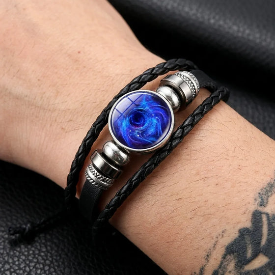 A close-up of a person's wrist wearing a multi-strand black leather bracelet with a central oval blue galaxy resin cabochon and silver-tone metal beads.