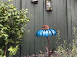 A decorative metal flower garden stake shaped like a blue coneflower with a brown textured center, set in a garden against a dark green wooden fence with plants.