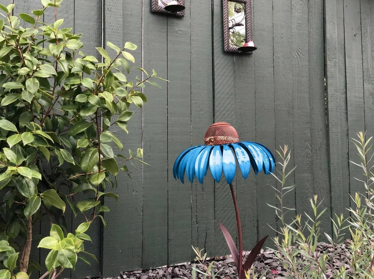 A decorative metal flower garden stake shaped like a blue coneflower with a brown textured center, set in a garden against a dark green wooden fence with plants.