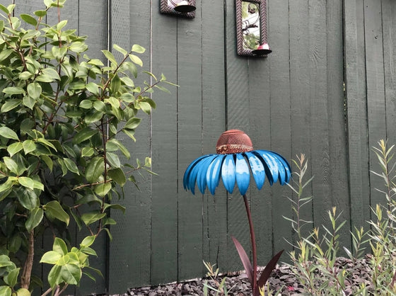 A decorative metal flower garden stake shaped like a blue coneflower with a brown textured center, set in a garden against a dark green wooden fence with plants.