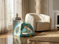 A small children's play tent with a colorful floral and geometric pattern in blue, yellow, and white, set up on a wooden floor in a bright, modern living room next to a cream boucle armchair.
