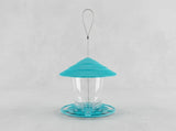 A bird feeder with a bright turquoise blue conical lid, a clear plastic cup in the center, and a matching turquoise base, suspended by a thin metal wire loop against a light gray background.