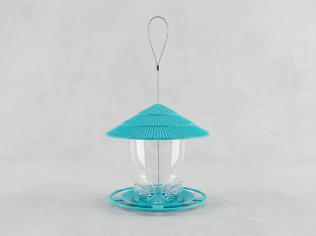 A bird feeder with a bright turquoise blue conical lid, a clear plastic cup in the center, and a matching turquoise base, suspended by a thin metal wire loop against a light gray background.