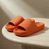 A pair of bright orange slide sandals with a ribbed strap and thick textured sole, displayed on a light beige stone table against a neutral beige background.