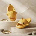 Two yellow shark-shaped slide sandals displayed on beige stone pedestals against a softly textured beige background, with small decorative leaves and stones around them.