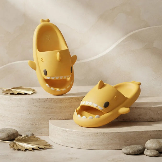 Two yellow shark-shaped slide sandals displayed on beige stone pedestals against a softly textured beige background, with small decorative leaves and stones around them.