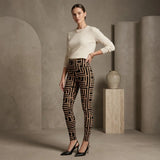 A woman models high-waisted leggings with a black and beige geometric maze-like pattern, paired with a cream long-sleeve top and black heels, in a minimalist studio setting with concrete-textured walls and a ceramic vase.