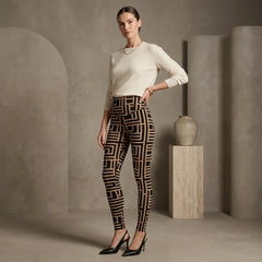 A woman models high-waisted leggings with a black and beige geometric maze-like pattern, paired with a cream long-sleeve top and black heels, in a minimalist studio setting with concrete-textured walls and a ceramic vase.