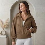 A woman wearing a brown quarter-zip mock-neck sweatshirt with a stand collar, paired with white pants, standing in front of a beige arched wall.