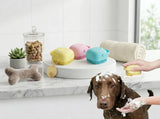 Three pet grooming brushes in yellow, pink, and blue are displayed on a white pedestal in a bright bathroom setting, with a dog covered in soap lather and a person's hand washing the dog. A rolled towel, bone-shaped toy, and pet treat jar are also visible.