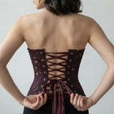 A woman shown from the back wearing a lace corset with burgundy laces, adjusting the back ties against a plain light gray background.