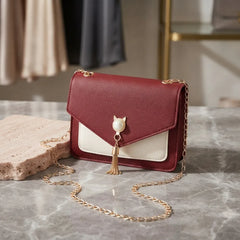 A small envelope-style shoulder bag in burgundy and cream faux leather with a gold chain strap, resting on a gray marble surface next to a stone block.